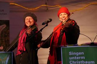 Monika Drasch (links) und Traudi Siferlinger (rechts) singen mit Geigen unterm Christbaum auf dem Münchner Christkindlmarkt
