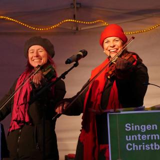 Monika Drasch (links) und Traudi Siferlinger (rechts) singen mit Geigen unterm Christbaum auf dem Münchner Christkindlmarkt